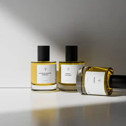 Three glass perfume bottles with black caps and white labels, filled with amber-yellow liquid, arranged on a white surface against a gray background. One bottle lies on its side.